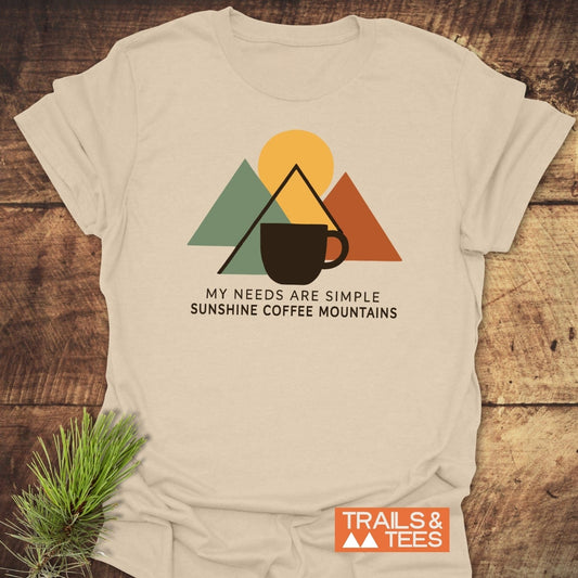 The Simple Joys T-Shirt features a mountain graphic with a sun and mug, laid on wood. Ideal for outdoor enthusiasts, made from soft, durable cotton.