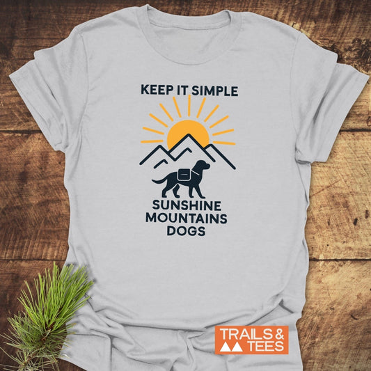 Minimalist Hiking Dog T-Shirt featuring a sunrise and mountains graphic, with a dog silhouette. Displayed on a wooden surface with a pine sprig beside it.