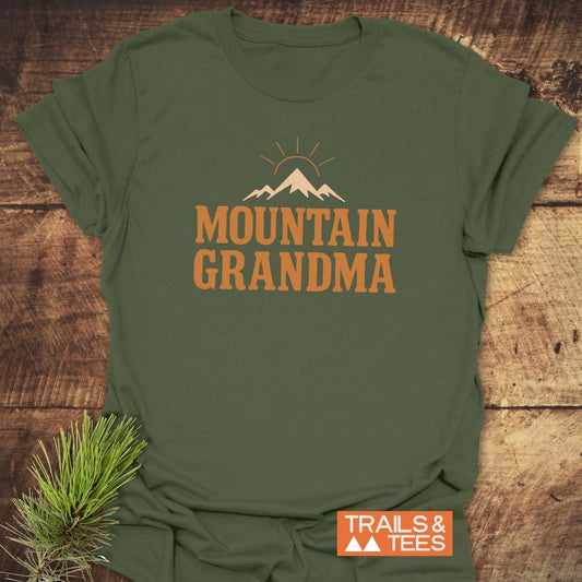 Mountain Grandma T-Shirt featuring a mountain silhouette and rising sun graphic, on a weathered wooden surface. Includes TRAILS & TEES label with pine needle accent.