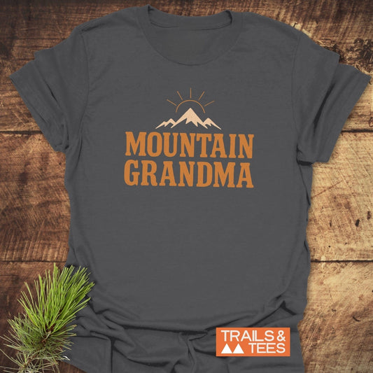 Mountain Grandma T-Shirt featuring a mountain peak and rising sun graphic, laid on wood. Made of soft, ring-spun cotton for comfort.