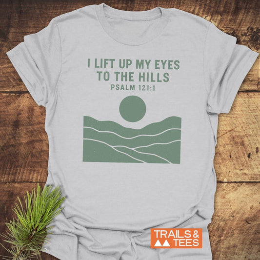 I Lift My Eyes... Mountain T-Shirt with green hills and sun graphic, Psalm 121:1 text, laid on wood, showcasing outdoor-inspired design and comfort.