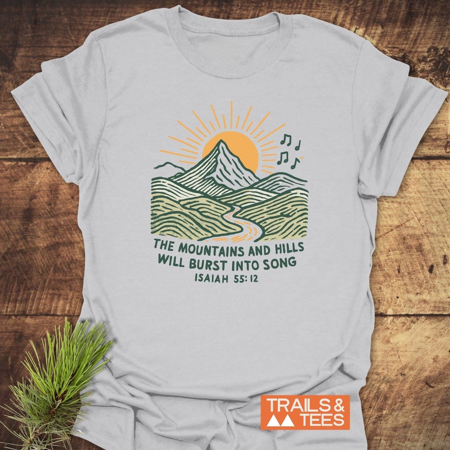 Mountains And Hills T-Shirt with a sun, mountain, and musical notes graphic. Includes Isaiah 55:12 quote. Features comfort-focused, durable design from Trails & Tees.