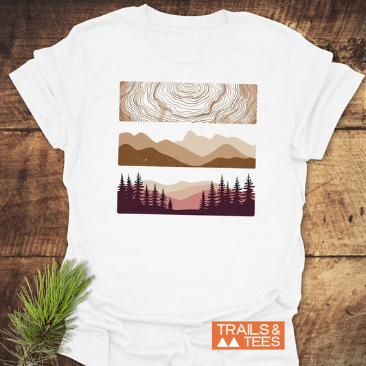 Minimalist Mountains Graphic T-Shirt featuring topographic lines, mountain silhouettes, and pine trees with TRAILS & TEES logo, laid on a wooden surface with pine sprig.