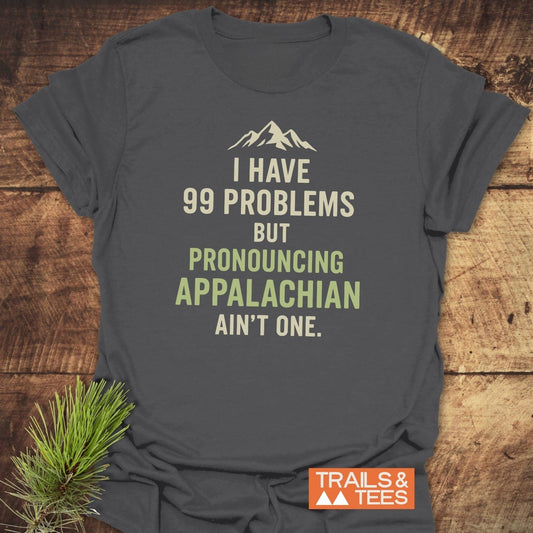 99 Problems Funny T-Shirt on wood surface, features mountain icon and humorous text about pronouncing Appalachian. Includes Trails & Tees logo in corner.