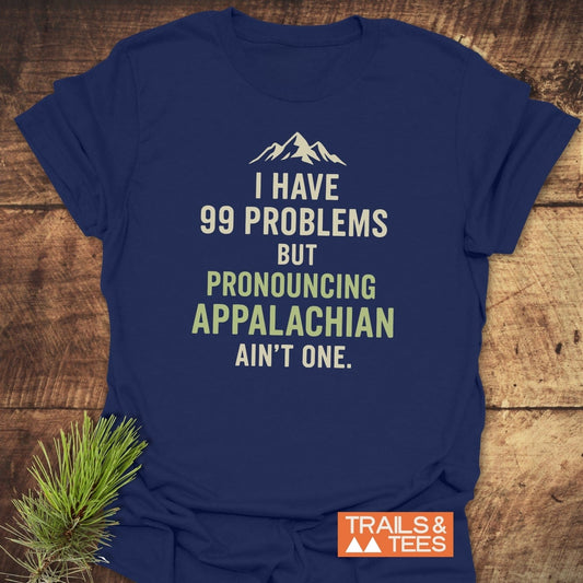 Navy-blue 99 Problems Funny T-Shirt with mountain graphic, playful slogan, and Trails & Tees logo, laid flat on a rustic surface with pine sprig.