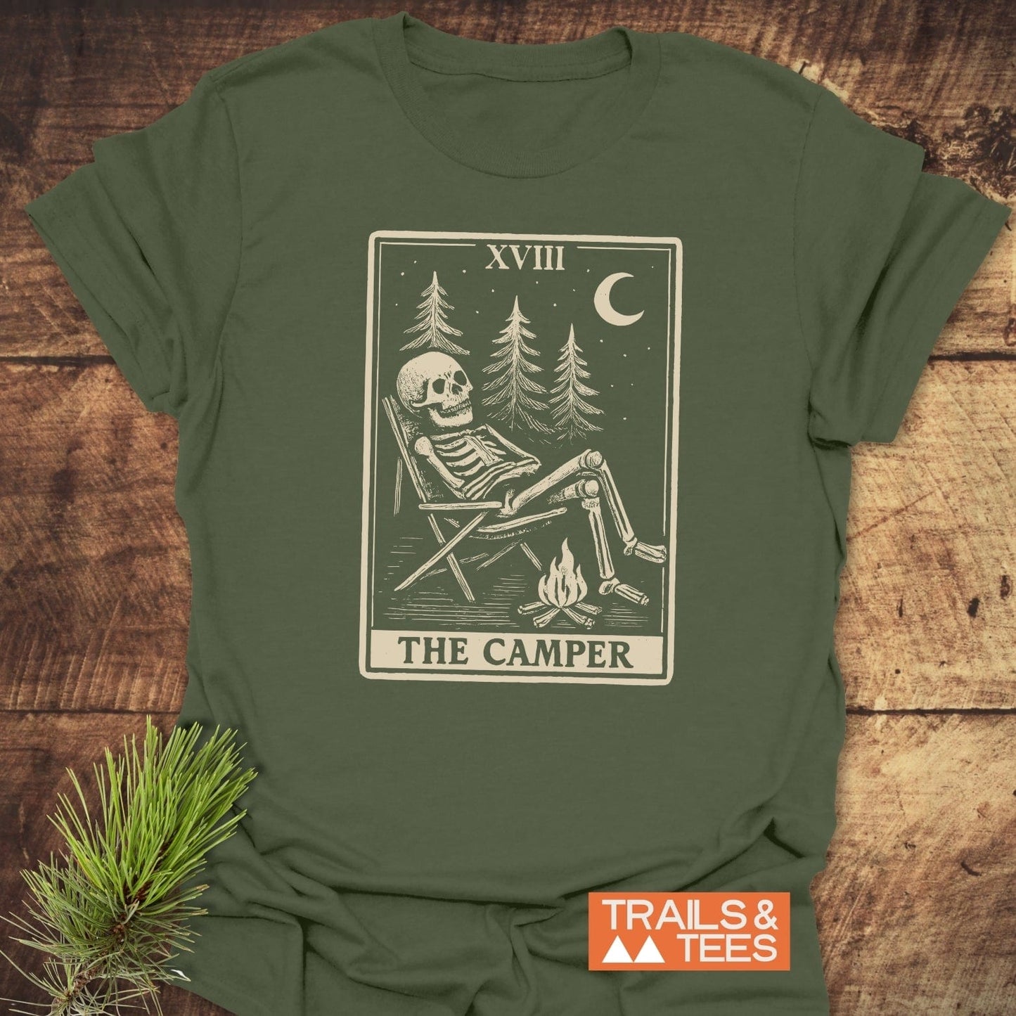 The Camper Tarot T-Shirt with skeleton in camp scene, pine trees, moon, and stars, showcasing outdoor adventure. Features durable print and comfy, pre-shrunk cotton.