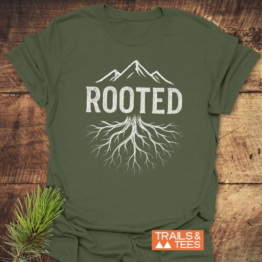 Olive-green Rooted Mountain T-Shirt featuring a white mountain graphic and ROOTED text with tree roots, laid on wood with a pine sprig.