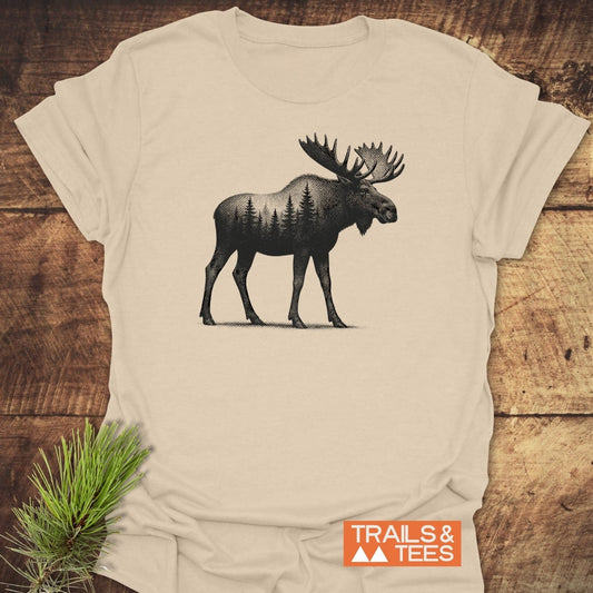 Moose Halftone T-Shirt featuring a moose with evergreen trees illustration on soft, lightweight cotton, laid on wood with TRAILS & TEES logo and pine sprig.