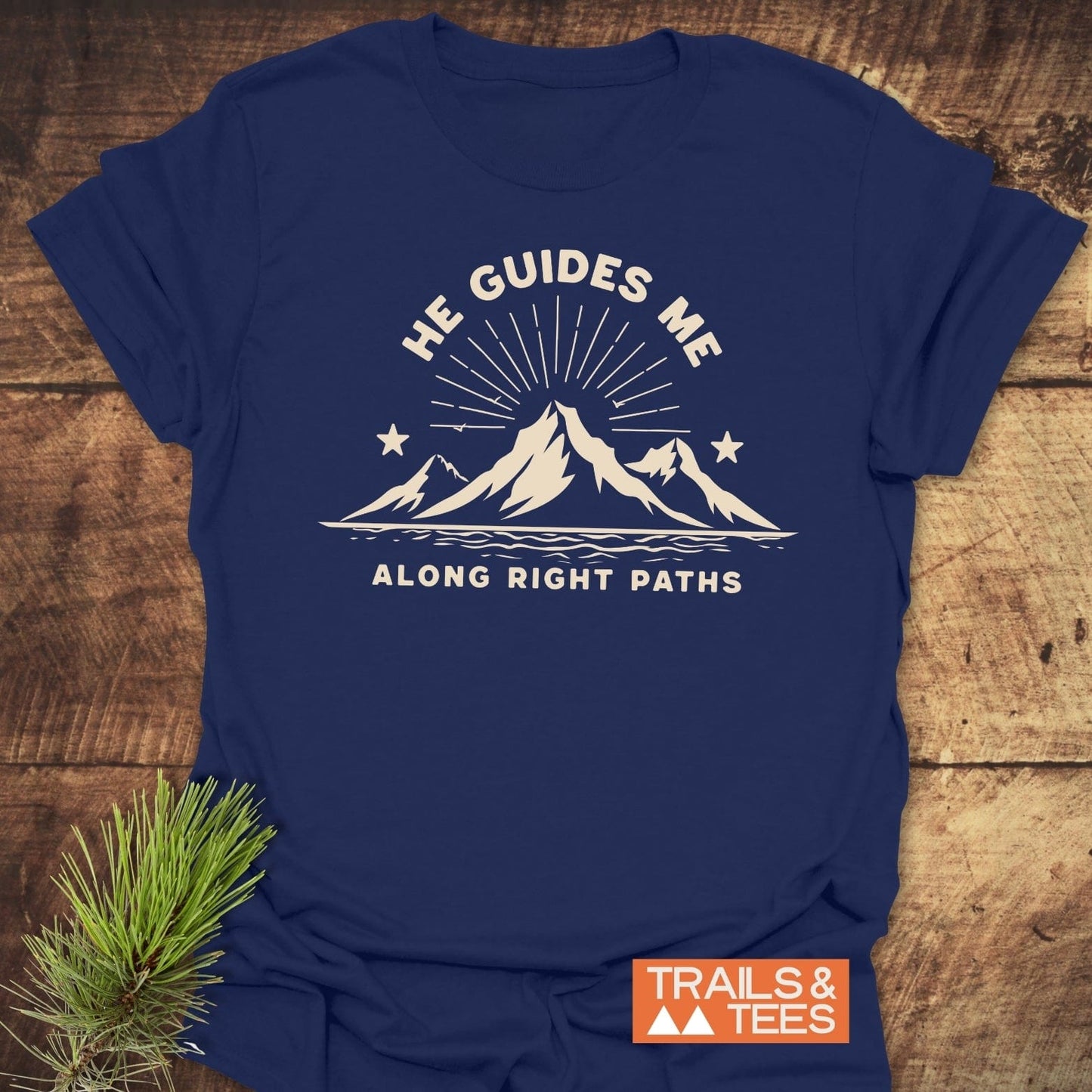 He Guides Me T-Shirt with mountain graphic, showcasing durable print and soft cotton, styled on wood with pine sprig, embodies outdoor adventure and comfort.