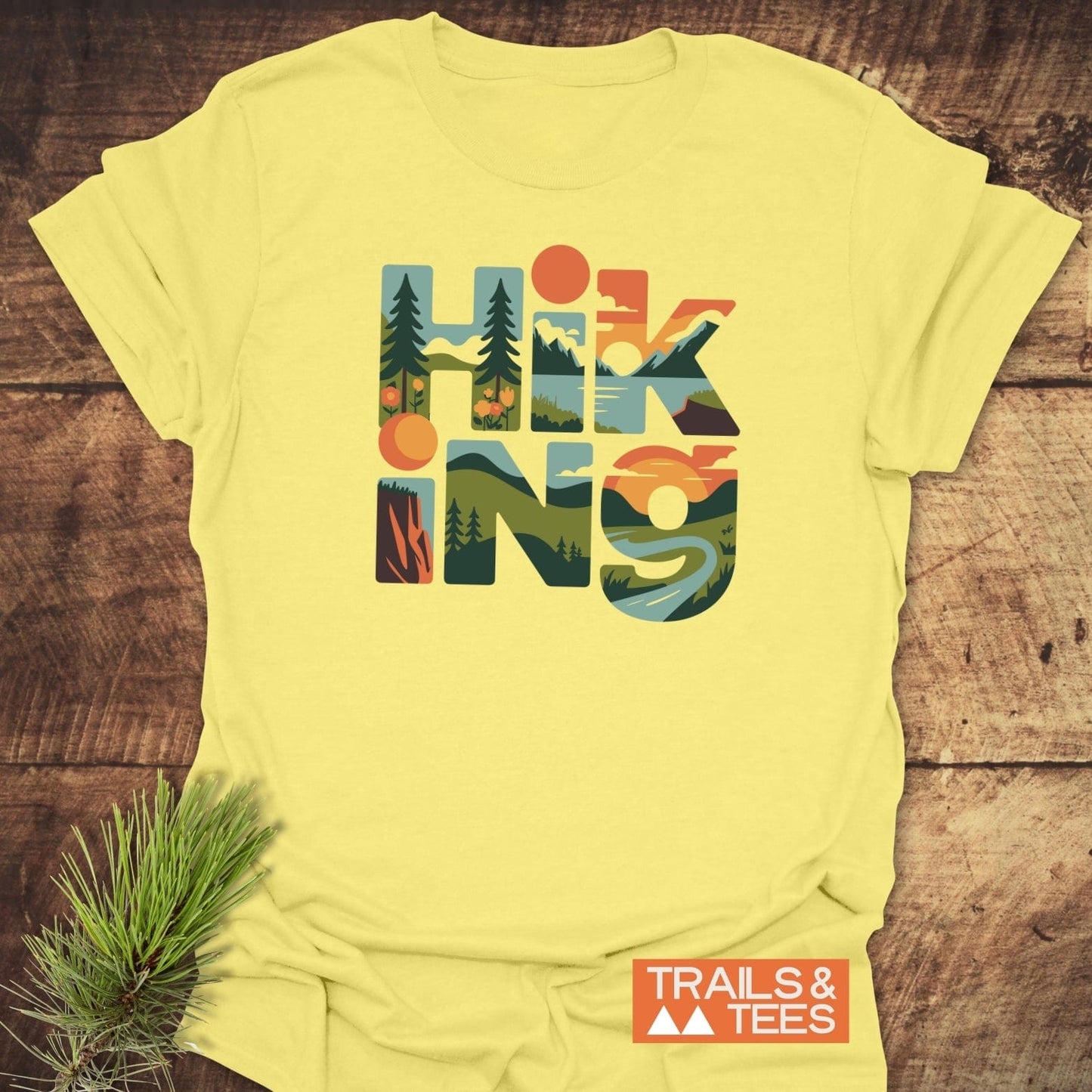 Hiking Typographic T-Shirt on rustic surface, featuring HIKING in outdoor-themed block letters, with TRAILS & TEES logo and pine sprig accentuating the nature theme.