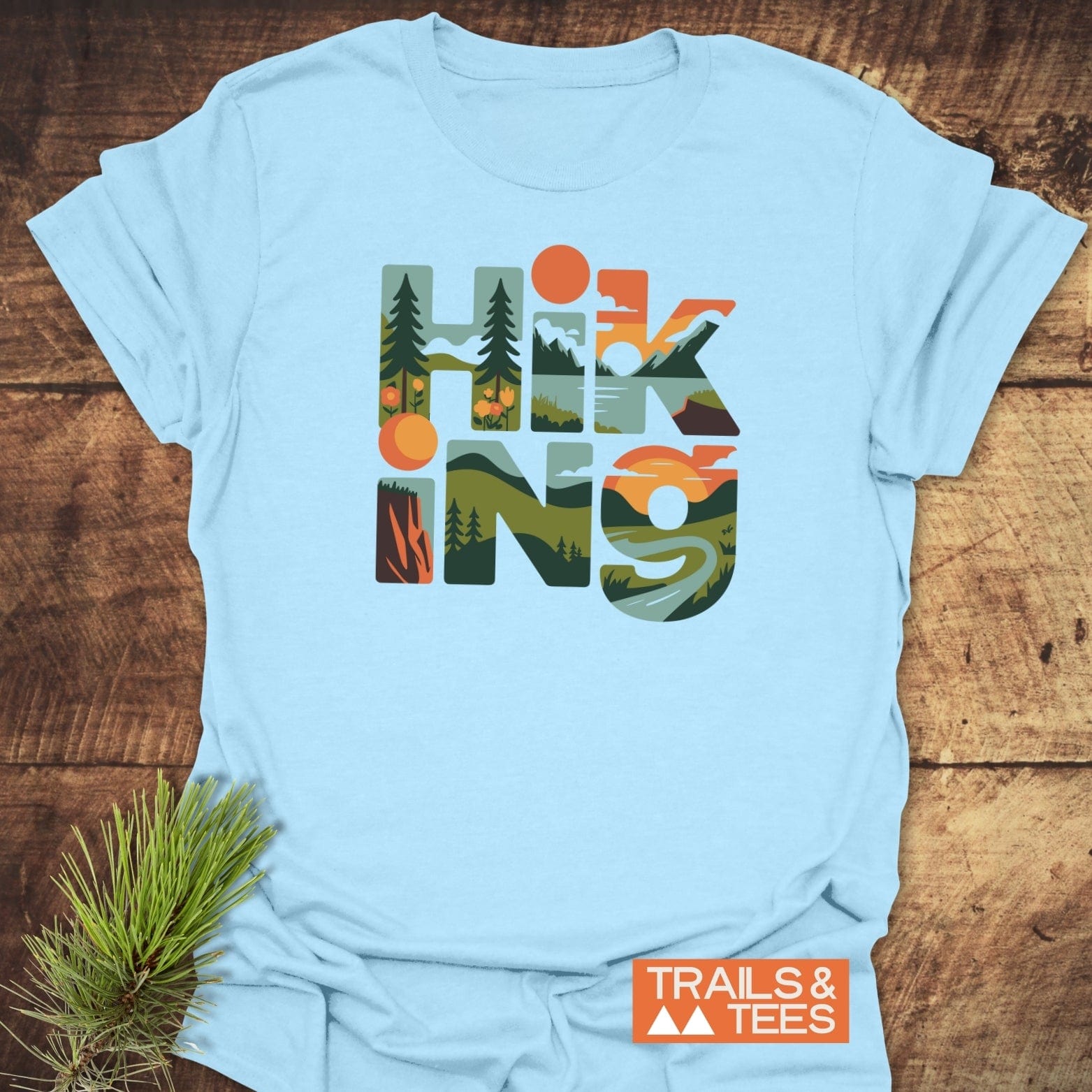 Hiking Typographic T-Shirt: Light blue tee with HIKING in nature-themed letters, featuring durable prints. Includes TRAILS & TEES logo and pine twig prop.
