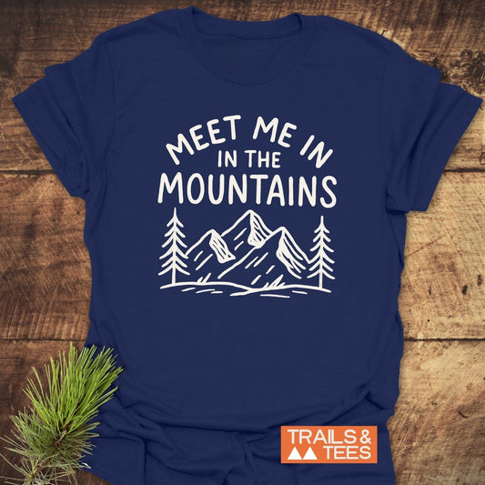Meet Me In The Mountains T-Shirt with hand-drawn mountain graphic, white print on navy, pine tree accents, and Trails & Tees label, emphasizing outdoor adventure.