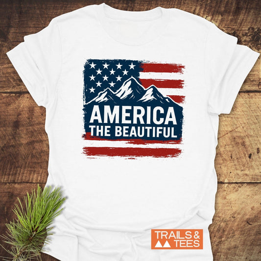 America The Beautiful T-Shirt featuring a distressed American flag with mountains and slogan, on soft cotton, laid on wood with Trails & Tees logo.