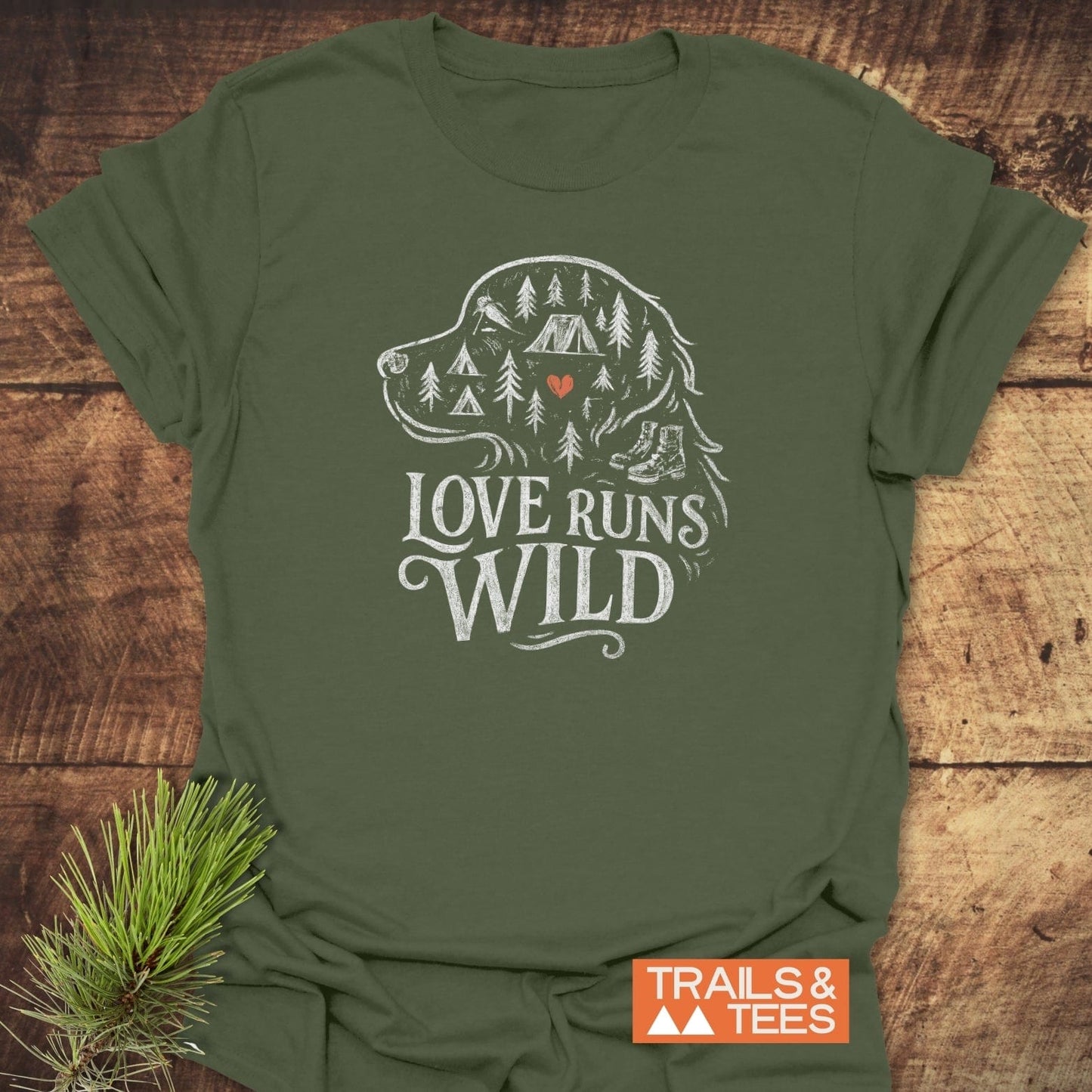 Love Runs Wild T-Shirt featuring an outlined dog graphic with camping motifs, rustic slogan, and Trails & Tees label, on a wooden surface with pine needles.