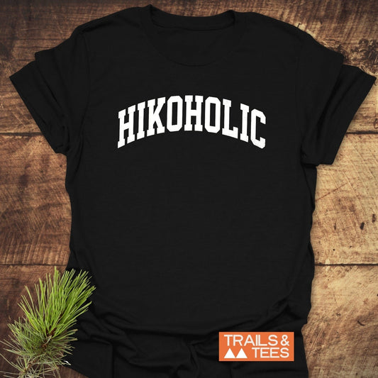 Hikoholic T-Shirt on wooden surface with “HIKOHOLIC” in white letters; pine sprig suggests adventure. Made of soft cotton, designed for outdoor enthusiasts.