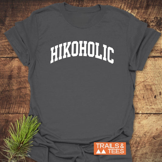 Charcoal-gray Hikoholic T-shirt with white block letters, laid flat. Features orange TRAILS & TEES logo and evergreen sprig for styling.