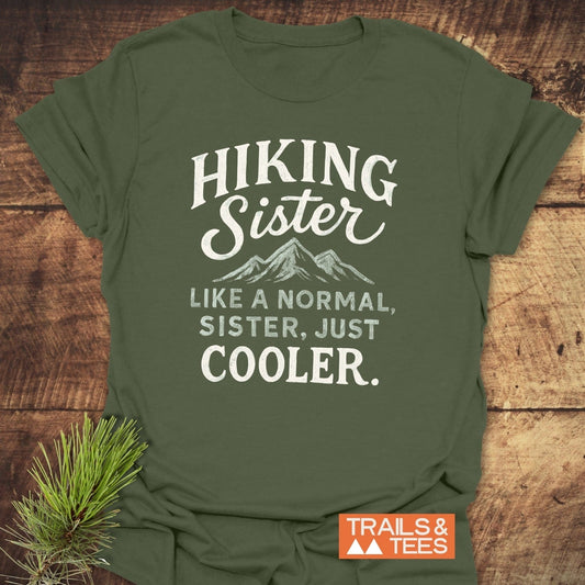 Olive-green Hiking Sister T-Shirt with white hand-lettered graphic, mountain peaks, and TRAILS & TEES logo, styled with a pine twig.