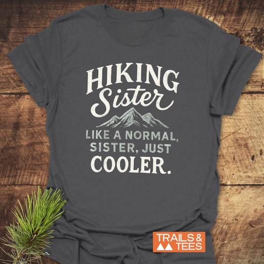 Charcoal-gray Hiking Sister T-Shirt with mountain graphic and slogan, laid on wood. Features durable print, soft cotton, pre-shrunk fit, styled with pine sprig.