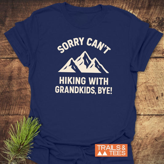 Navy-blue Sorry Hiking With Grandkids T-shirt with mountain graphic, laid on wooden surface, pine sprig beside. Features durable print and soft, lightweight cotton.
