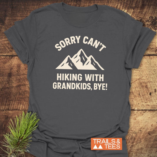 Sorry Hiking With Grandkids T-Shirt featuring white mountain graphic on dark-gray fabric, showcasing durable, comfortable design with TRAILS & TEES logo and pine sprig accent.