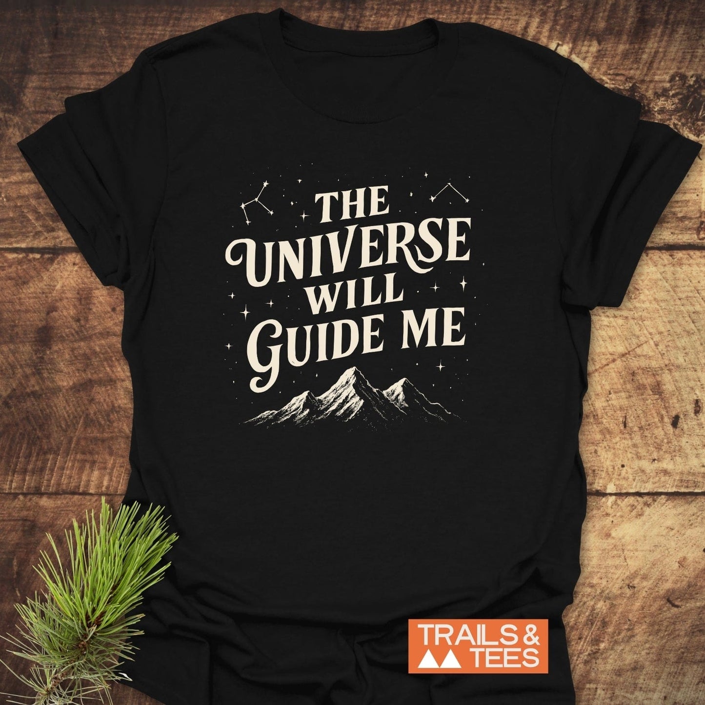 The Universe Guides Me T-Shirt featuring cream lettering and celestial designs on black cotton, styled with a pine sprig, embodies trail-ready comfort and outdoor spirit.