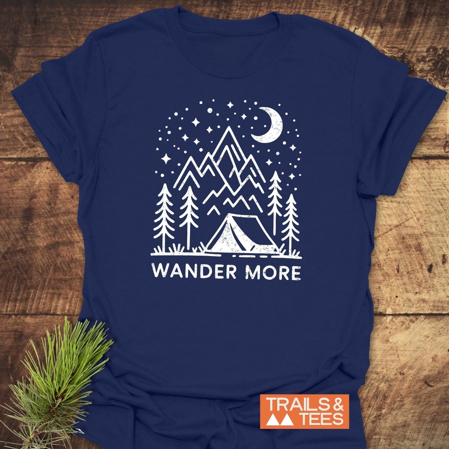 Navy-blue Wander More T-shirt with white line-art of a camping scene, featuring a tent, pine trees, mountains, and a starry sky.