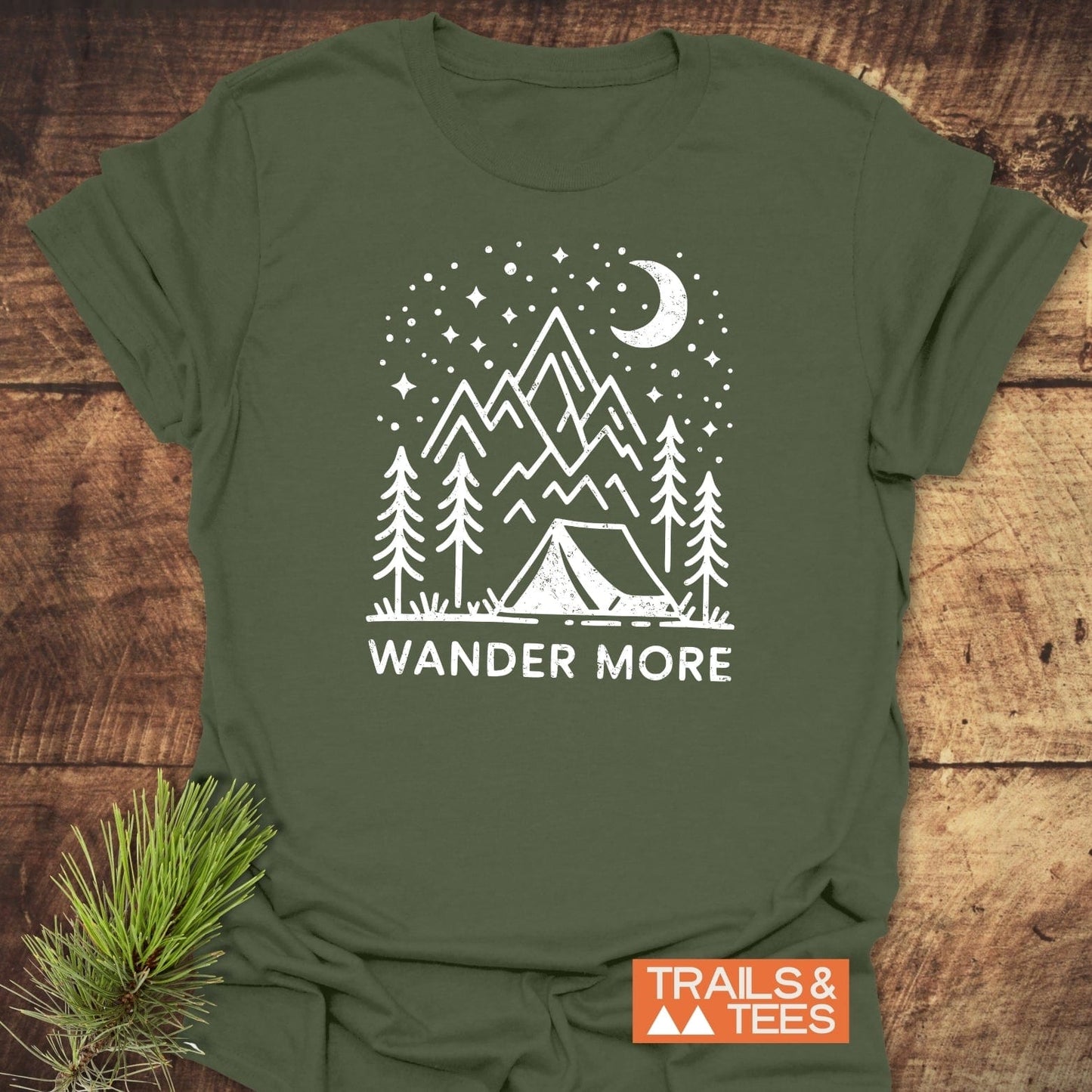Wander More T-Shirt featuring a hand-drawn camping scene with mountains, crescent moon, and stars. Includes TRAILS & TEES label, crafted for everyday adventure.