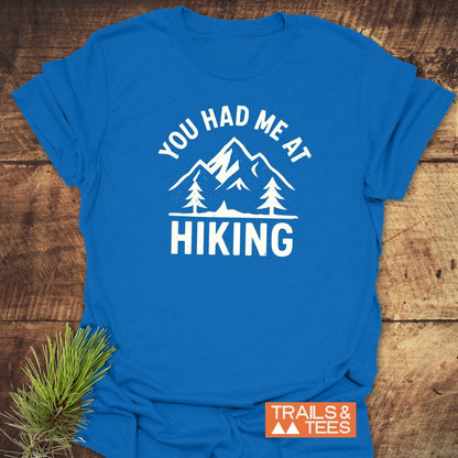 You Had Me At Hiking T-Shirt featuring a mountain range and pine trees design, emphasizing outdoor adventure. Displayed with Trails & Tees logo and spruce garnish.