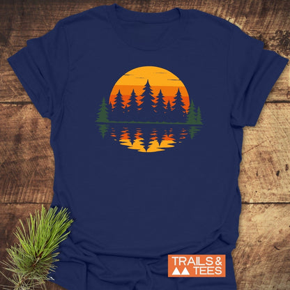 Retro Sunset Pines T-Shirt on a wood surface, features a sunset and evergreen design. Crafted from soft cotton, ideal for outdoor enthusiasts. Includes TRAILS & TEES logo.