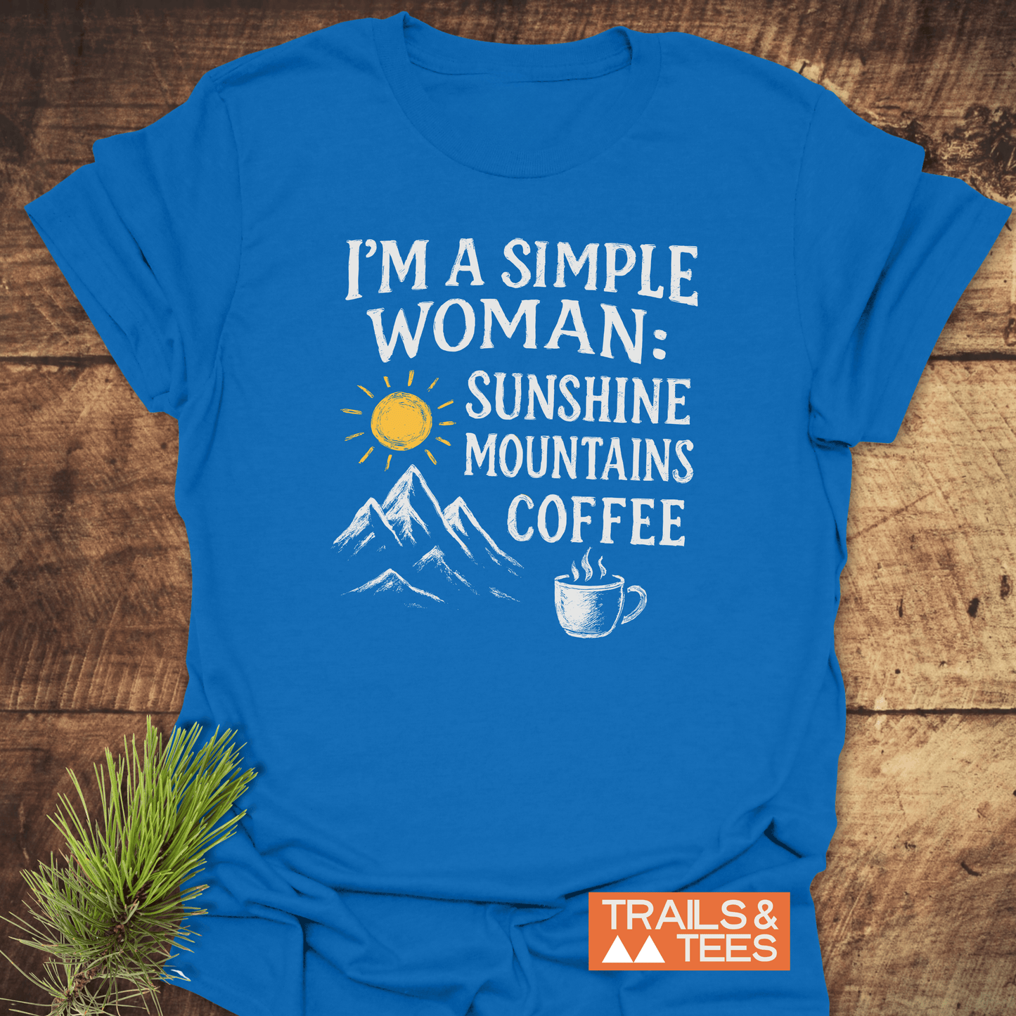 Simple Woman Sunshine T-Shirt flat-lay on wood, featuring sun, mountains, coffee mug prints. Includes TRAILS & TEES logo; crafted for adventure, pre-shrunk, durable design.