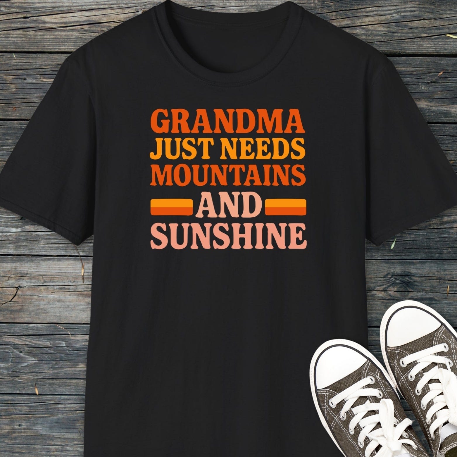 Retro 80s Grandma Mountains T-Shirt on wooden surface, with gray canvas sneakers. Bold, retro lettering celebrates love for mountains and sunshine.