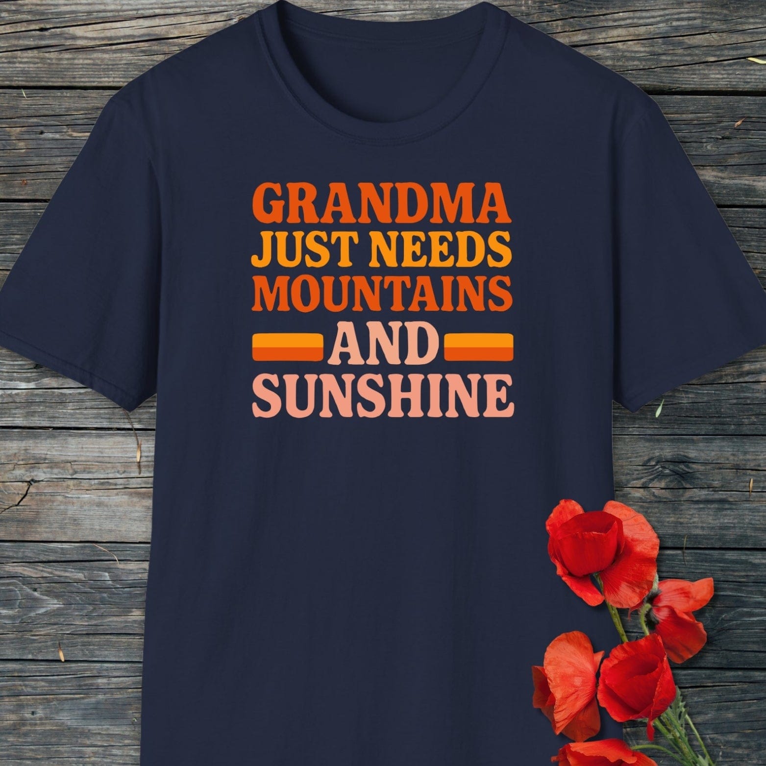 Retro 80s Grandma Mountains T-Shirt with bold, colorful slogan on soft navy fabric, laid flat; features bright red poppies beside it on a wooden surface.
