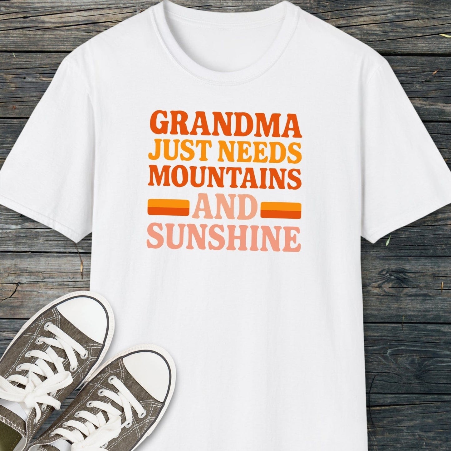 Retro 80s Grandma Mountains T-Shirt with bold slogan, laid on wood beside sneakers; crafted from soft cotton for comfort and adventure-ready durability.