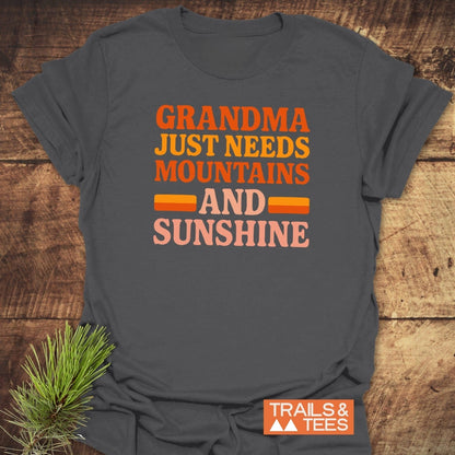 Retro 80s Grandma Mountains T-Shirt on wooden surface, featuring warm retro colors slogan, evergreen sprig, and TRAILS & TEES logo.
