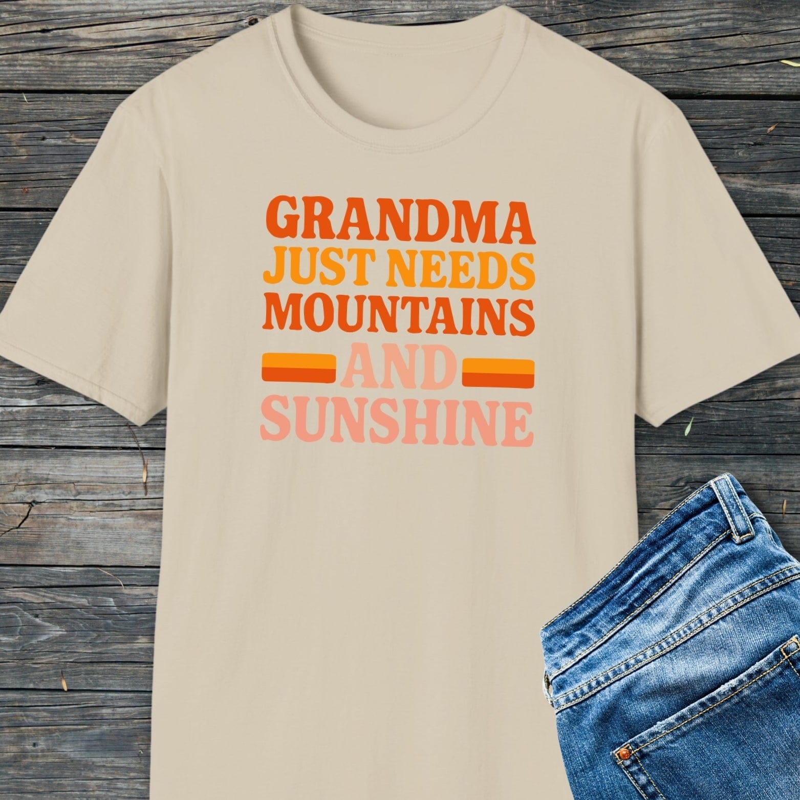 Retro 80s Grandma Mountains T-Shirt with colorful slogan, laid flat beside blue jeans on wooden surface, highlighting its soft, lightweight ring-spun cotton fabric.