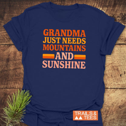 Retro 80s Grandma Mountains T-Shirt on wood surface, navy-blue with Grandma Just Needs Mountains And Sunshine print, beside pine sprig, Trails & Tees logo visible.