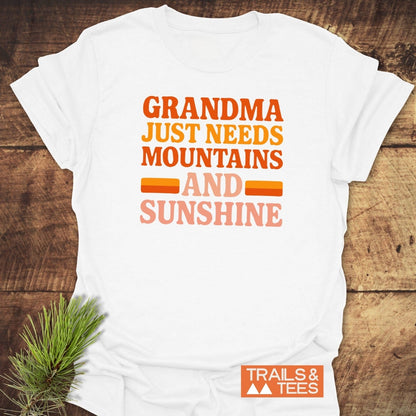 Retro 80s Grandma Mountains T-Shirt on rustic wood, featuring a vibrant slogan in bold lettering. Includes Trails & Tees logo and styled pine bough.
