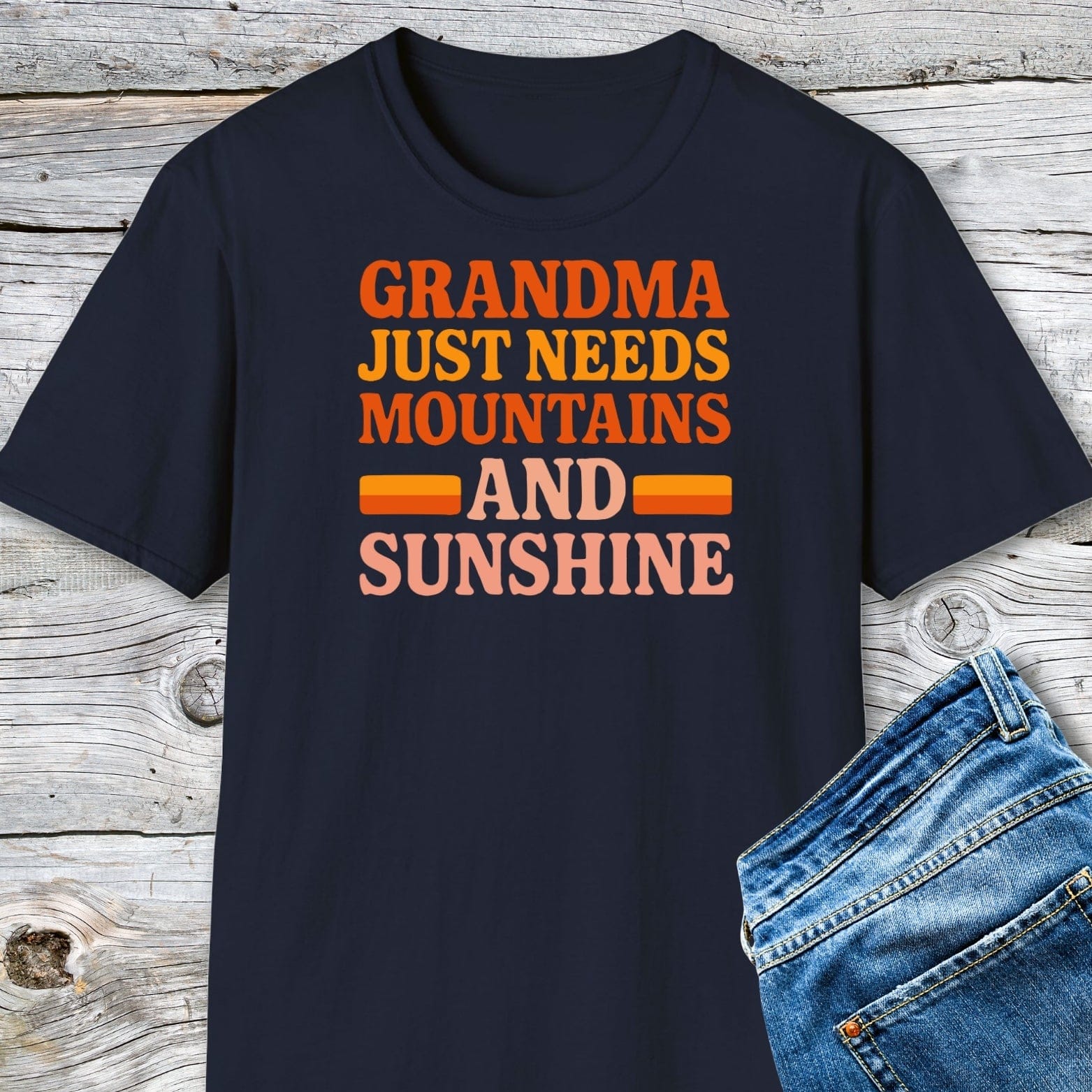 Retro 80s Grandma Mountains T-Shirt with multicolored slogan, laid flat on wood surface. Soft, durable ring-spun cotton, ideal for outdoor enthusiasts.