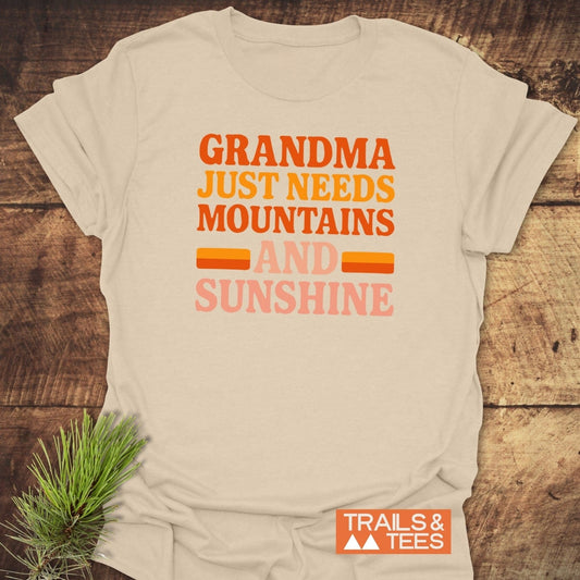 Retro 80s Grandma Mountains T-Shirt on wooden surface, featuring multicolored slogan. Includes small TRAILS & TEES logo. Pine sprig accentuates outdoorsy theme.