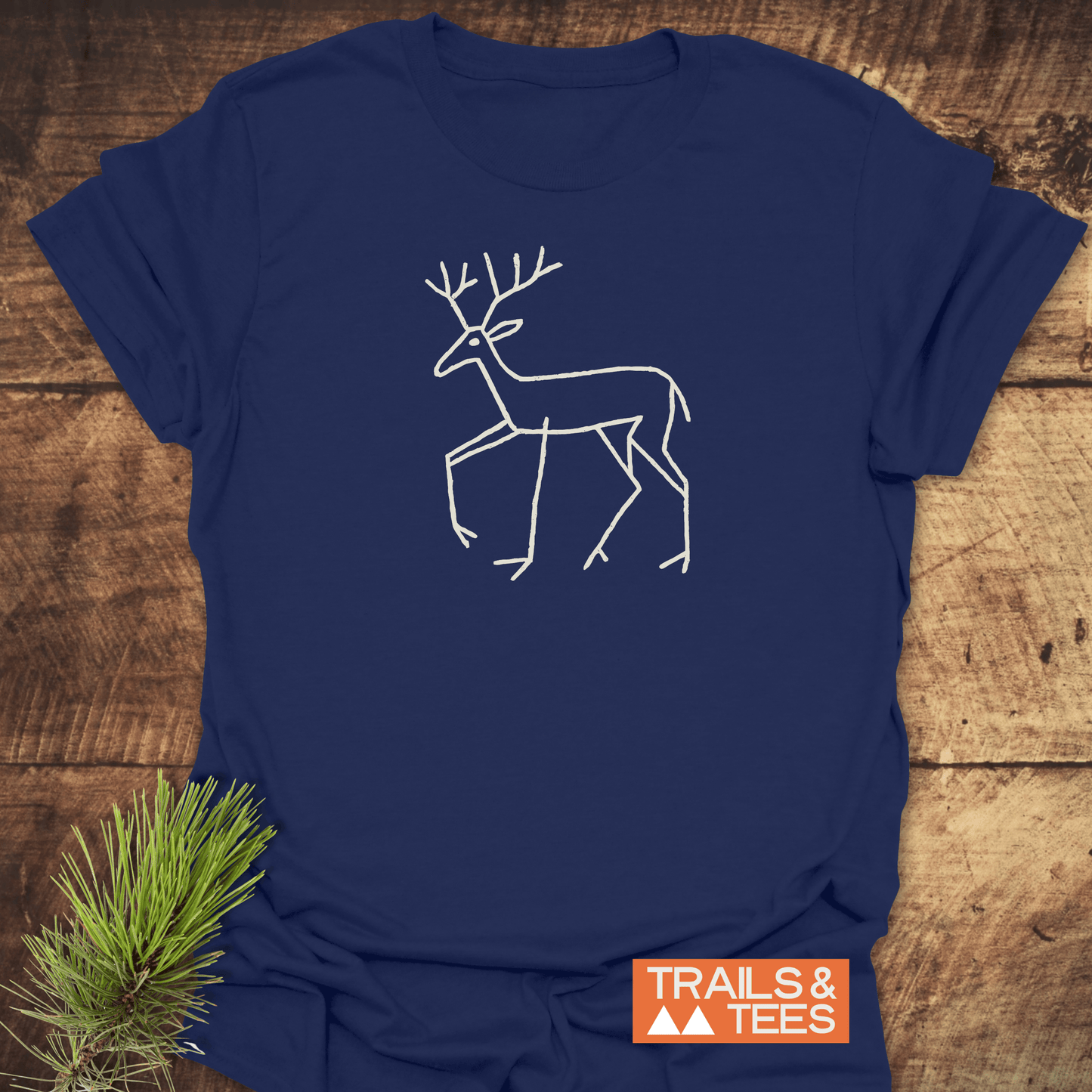 Deer Tribal T-Shirt featuring a white line-drawn stag design on navy-blue fabric, laid on wood with pine sprig, and TRAILS & TEES logo visible.