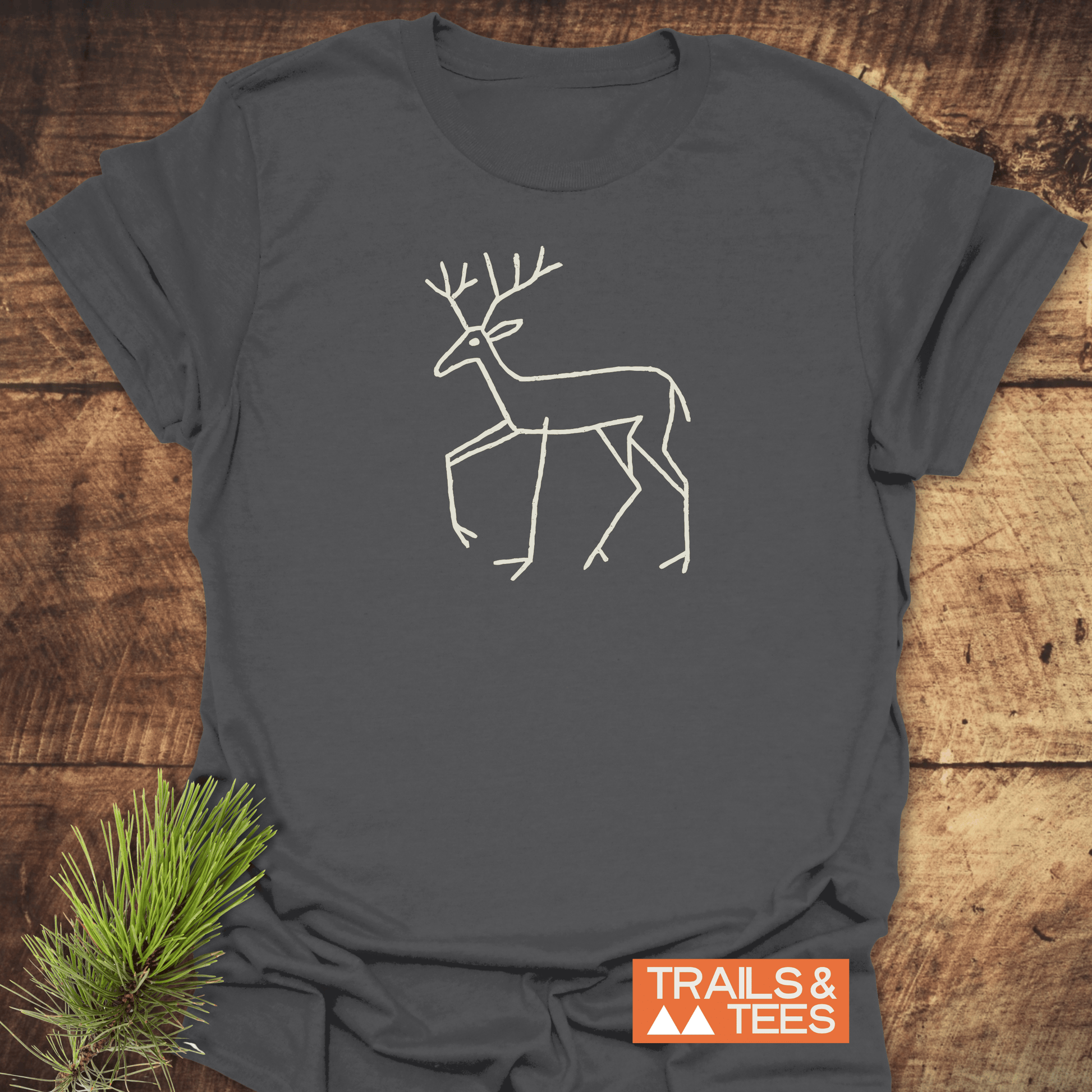 Deer Tribal T-Shirt featuring a line drawing of a deer with antlers, showcasing petroglyph-style design, on a charcoal-gray fabric. Includes TRAILS & TEES logo.