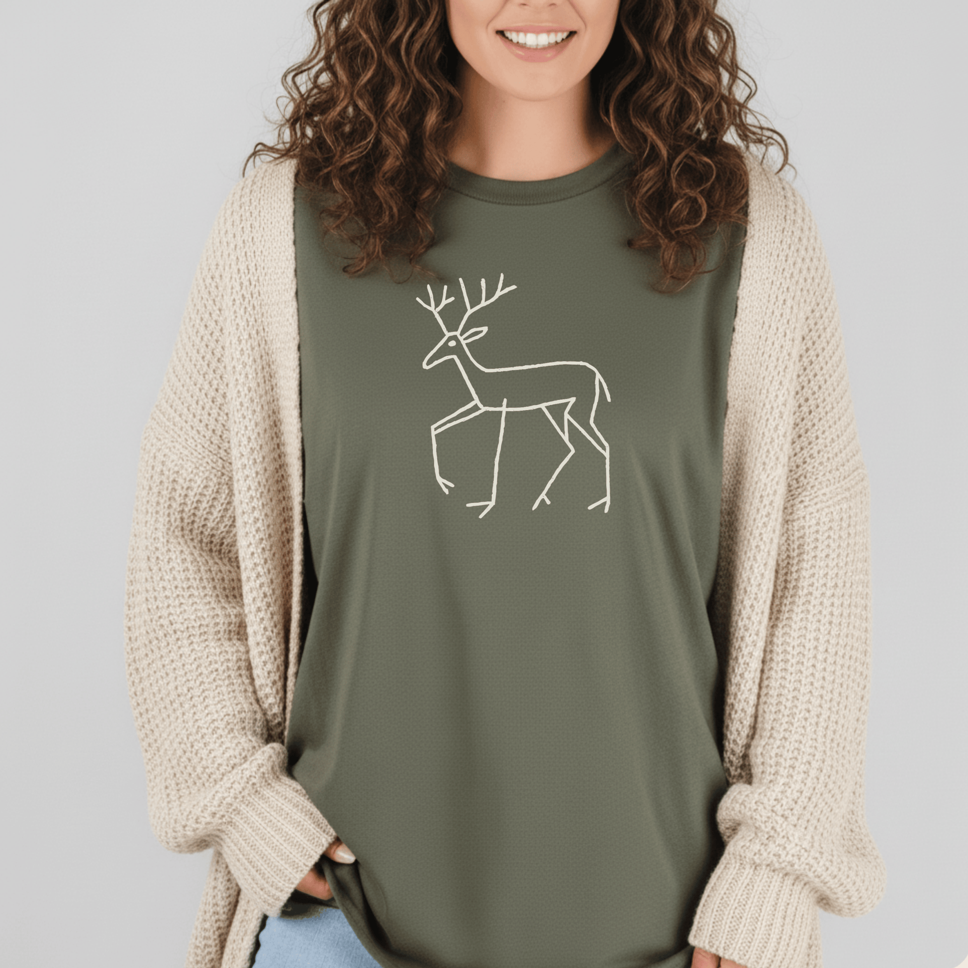 Person wearing Deer Tribal T-Shirt with a deer line drawing, layered with a chunky-knit cardigan, and blue jeans peeking at the bottom.