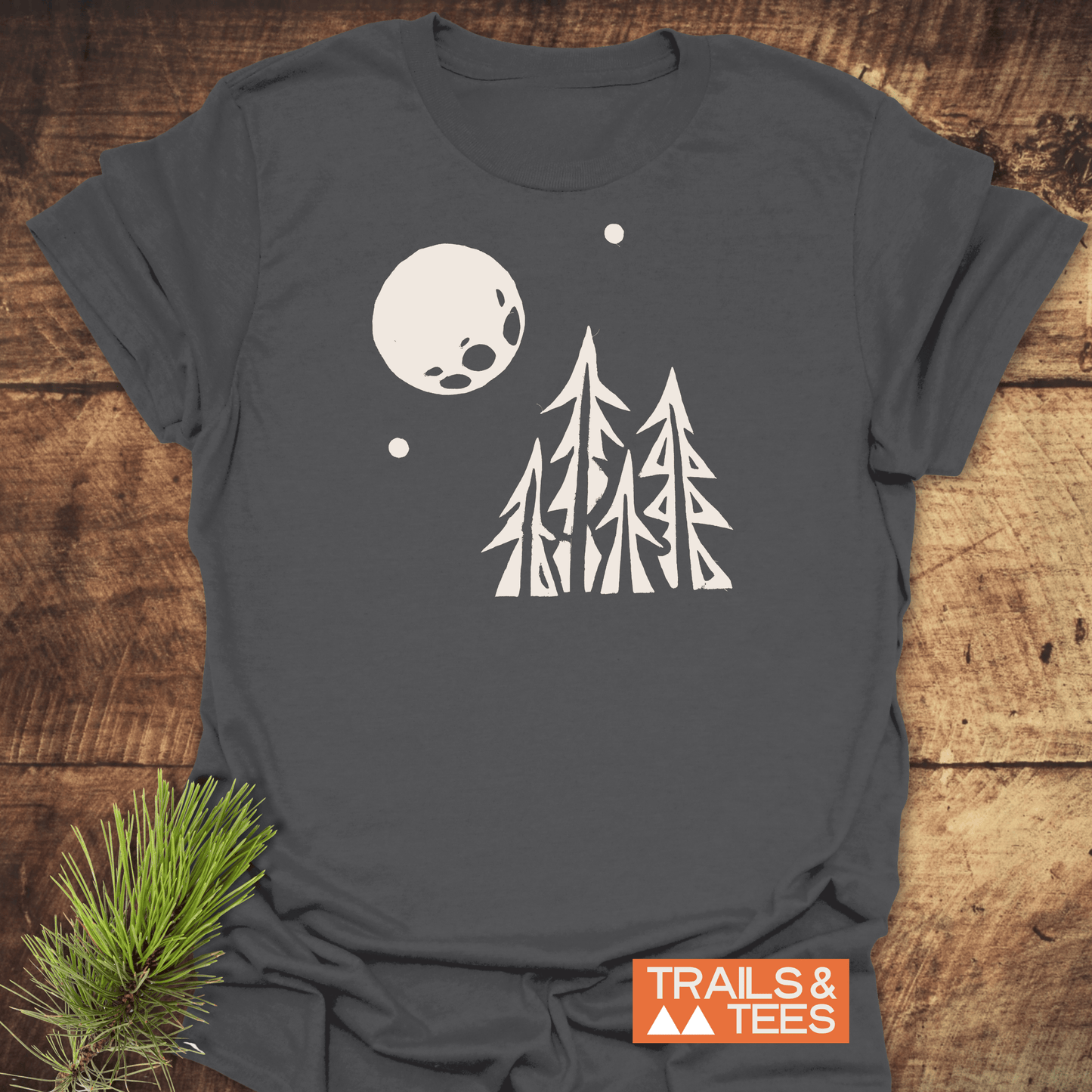 Pine Trees and Full Moon T-Shirt displayed on wooden surface, featuring white pine tree graphic under moon. Includes TRAILS & TEES logo, emphasizes outdoor adventure theme.
