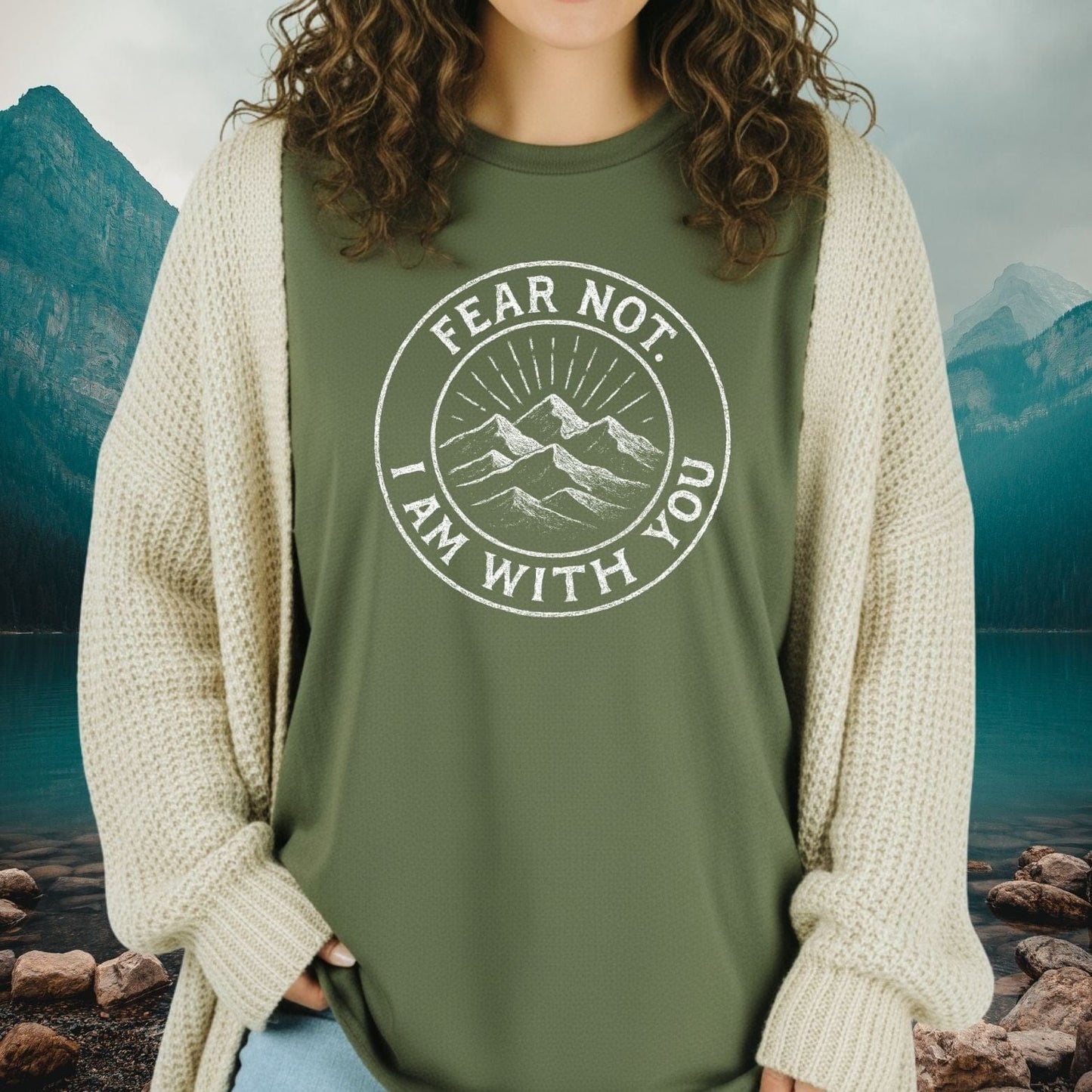 A person wears the Fear Not Mountain T-Shirt, featuring a circular mountain graphic, under a chunky cardigan, set against a serene mountain-lake backdrop.