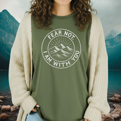 A person wears the Fear Not Mountain T-Shirt, featuring a circular mountain graphic, under a chunky cardigan, set against a serene mountain-lake backdrop.