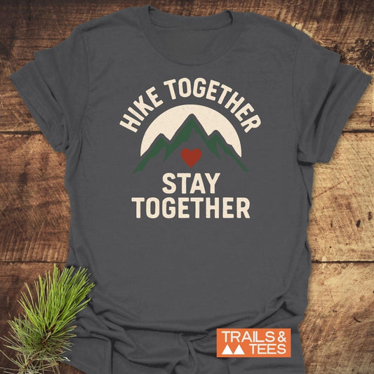 Hike Together Couple T-Shirt on wood, featuring a mountain graphic with heart, “HIKE TOGETHER, STAY TOGETHER” text, and “TRAILS & TEES” label, surrounded by pine sprigs.