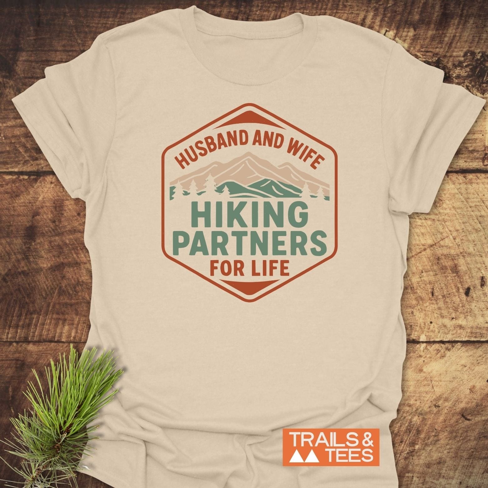 Hiking Partners For Life T-Shirt with mountain graphic and Husband and Wife text on soft, durable cotton, laid on rustic wood with pine sprig.