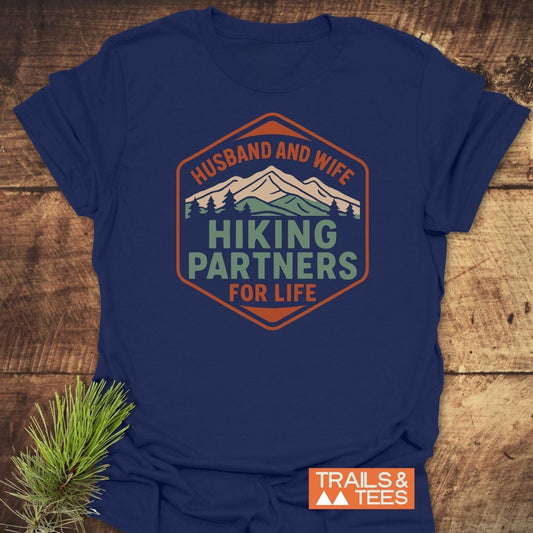 Navy-blue Hiking Partners For Life T-shirt featuring a mountain badge graphic, laid on wood, highlighting durable, soft cotton for outdoor enthusiasts.