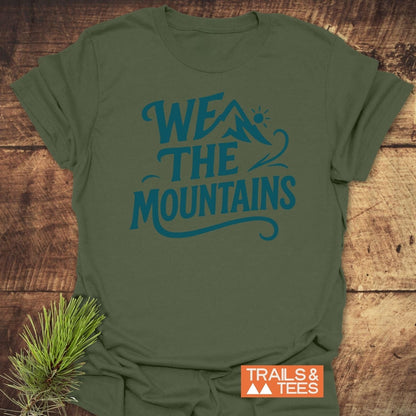 We The Mountains T-Shirt on wood surface; features mountain graphic and sun. Includes “TRAILS & TEES” logo and pine branch accent.