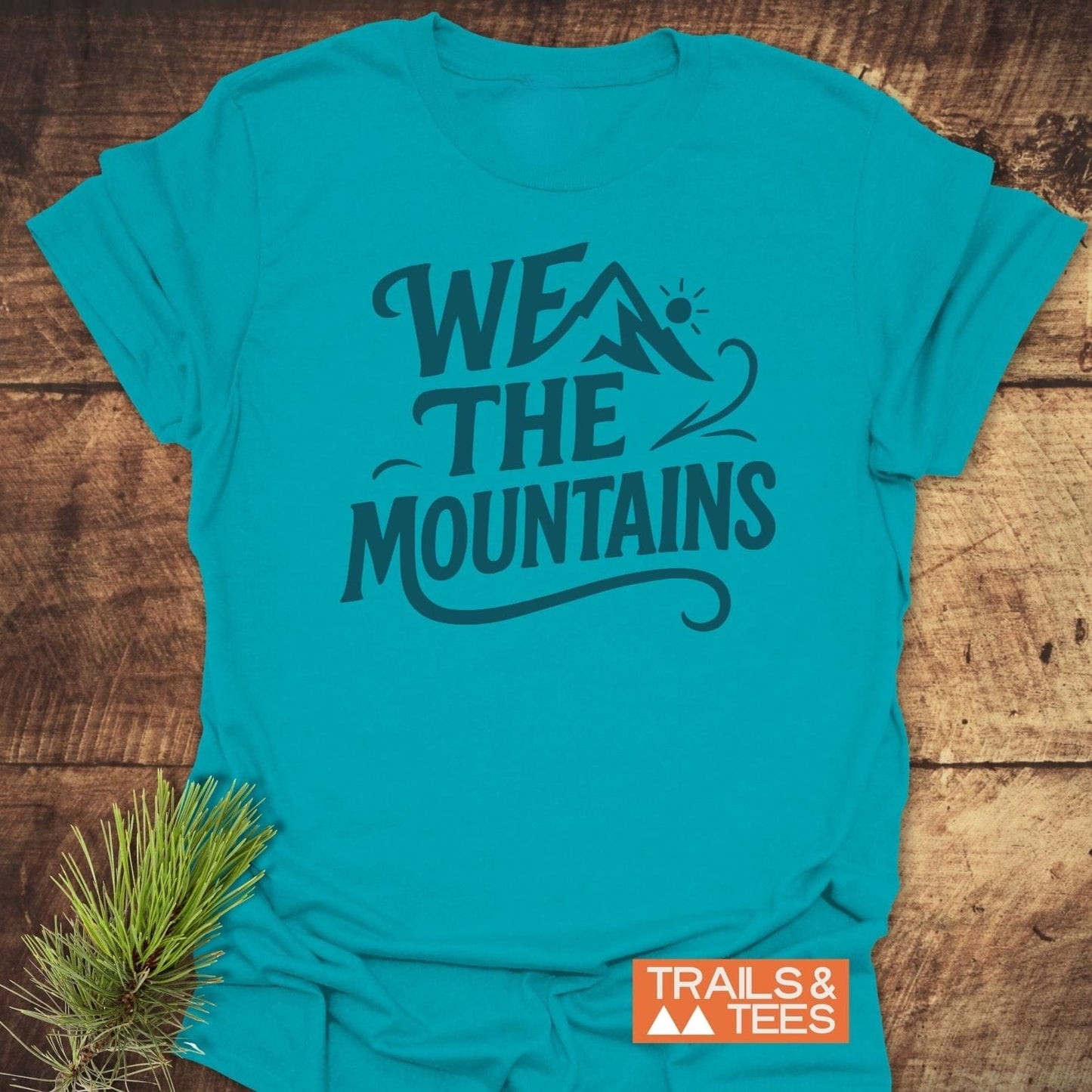 We The Mountains T-Shirt with mountain peak graphic, laid on wood. Soft, ring-spun cotton, pre-shrunk, durable print. Includes “TRAILS & TEES” logo and pine sprig.